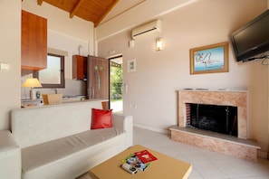 A living room with a fireplace, a sofa, a television, and a kitchen area visible through an open door. - Villa 'Ariadni' in Marathiás (Marathiás)