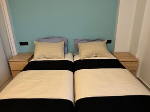 A twin room with two single beds, each with a pillow and a blanket. There are two bedside tables with lamps.