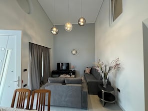 A modern living room with a grey sofa, a television, a small side table with a vase, and a dining area with a wooden table and chairs.