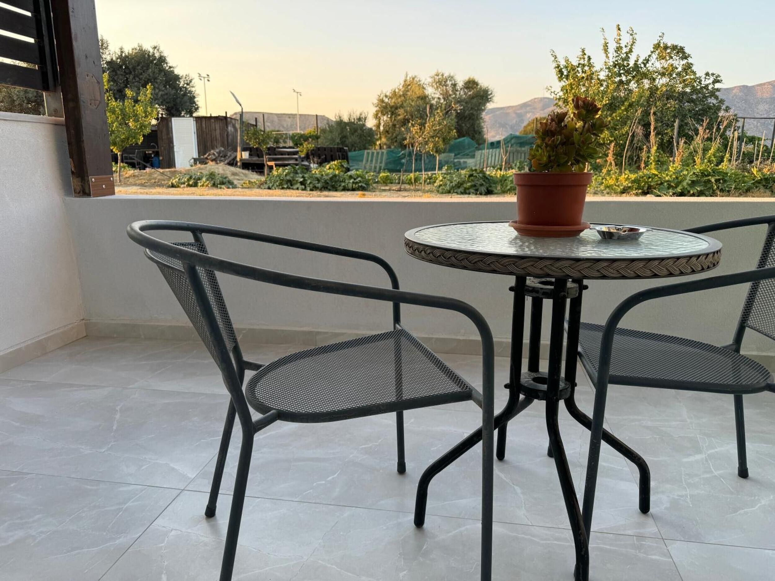 A terrace with two metal chairs and a table, a potted plant, and a view of a garden and mountains in the background.