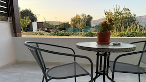 A terrace with two metal chairs and a table, a potted plant, and a view of a garden and mountains in the background.