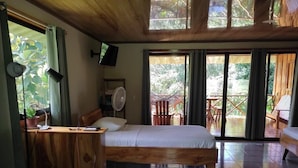 Ein Zimmer mit einem Bett, einem Schreibtisch mit Stuhl, einem Ventilator und einem Fernseher. Es gibt Schiebeglastüren zu einem Außenbereich mit Stühlen und einem Tisch.