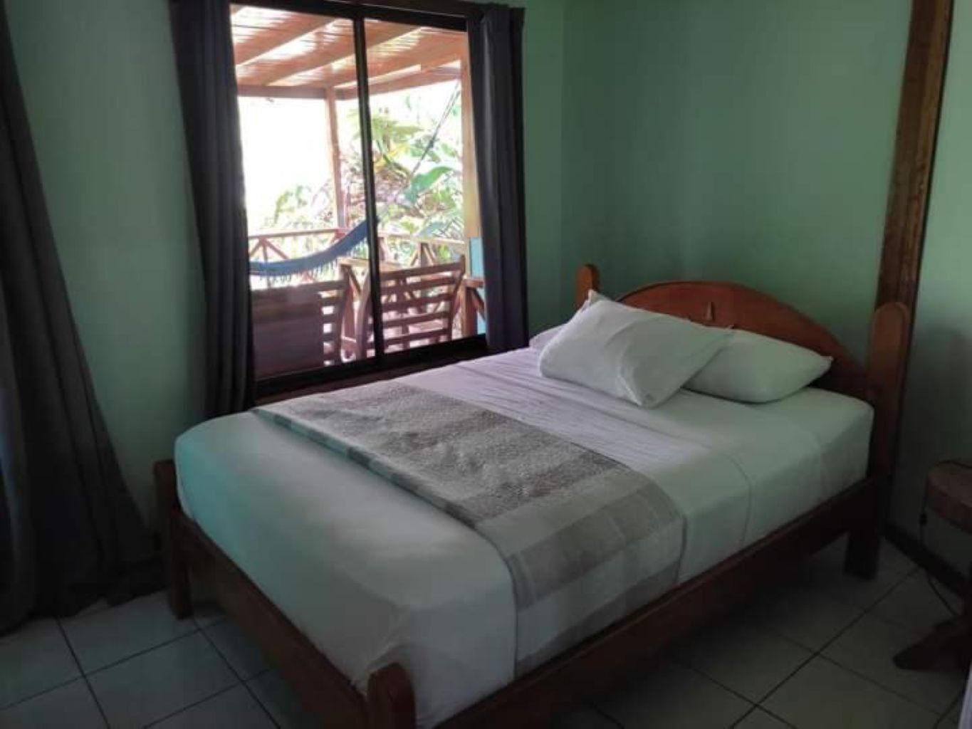 A single bed with a wooden headboard, a window with a view of outdoor furniture, and a tiled floor.