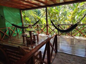 A wooden deck with a hammock, tables, and chairs, surrounded by lush greenery.