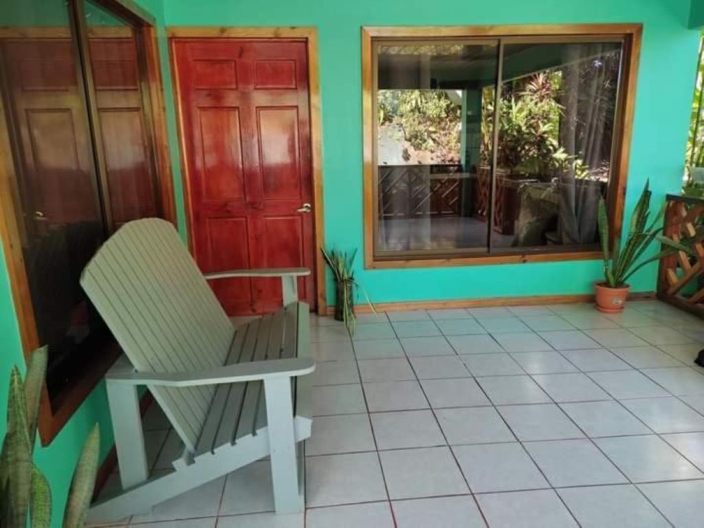 A room with a single chair, a red door, a window with a view of greenery, and a tiled floor.