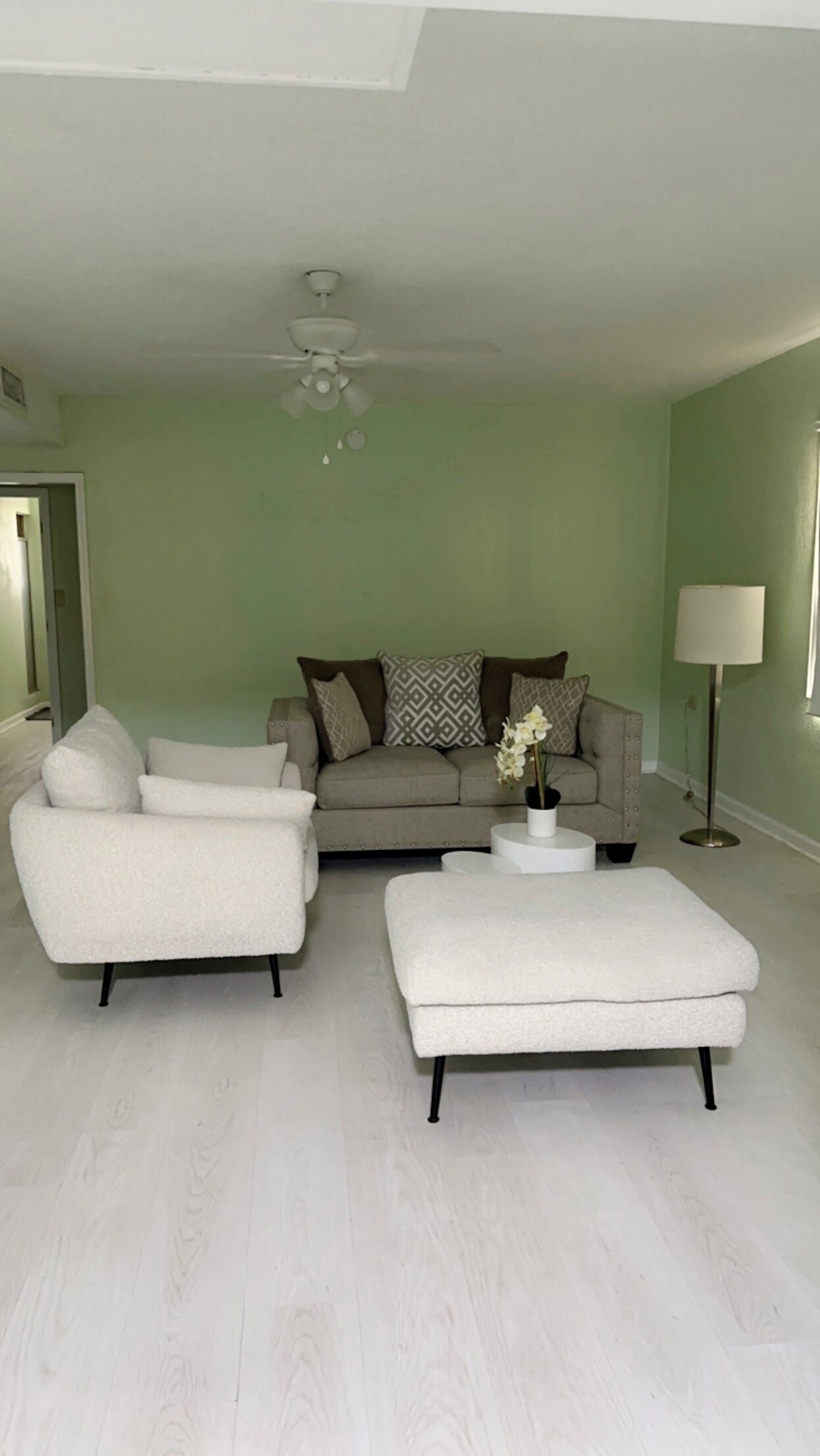 Un salon avec un canapé gris, des fauteuils blancs, un pouf blanc, une petite table d’appoint blanche et un lampadaire.