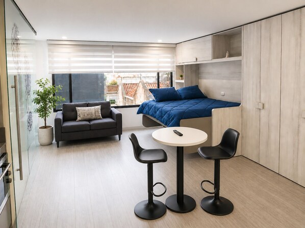 A modern, compact living space with a sofa, a small round table, and two bar stools. A bed with a blue cover is against the wall, and there is a potted plant on the floor. - Modern Studio in Chapinero | City Views & Workspace (Bogotá)