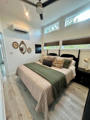 Een slaapkamer met een bed, nachtkastje, lamp, wandairconditioner en plafondventilator.