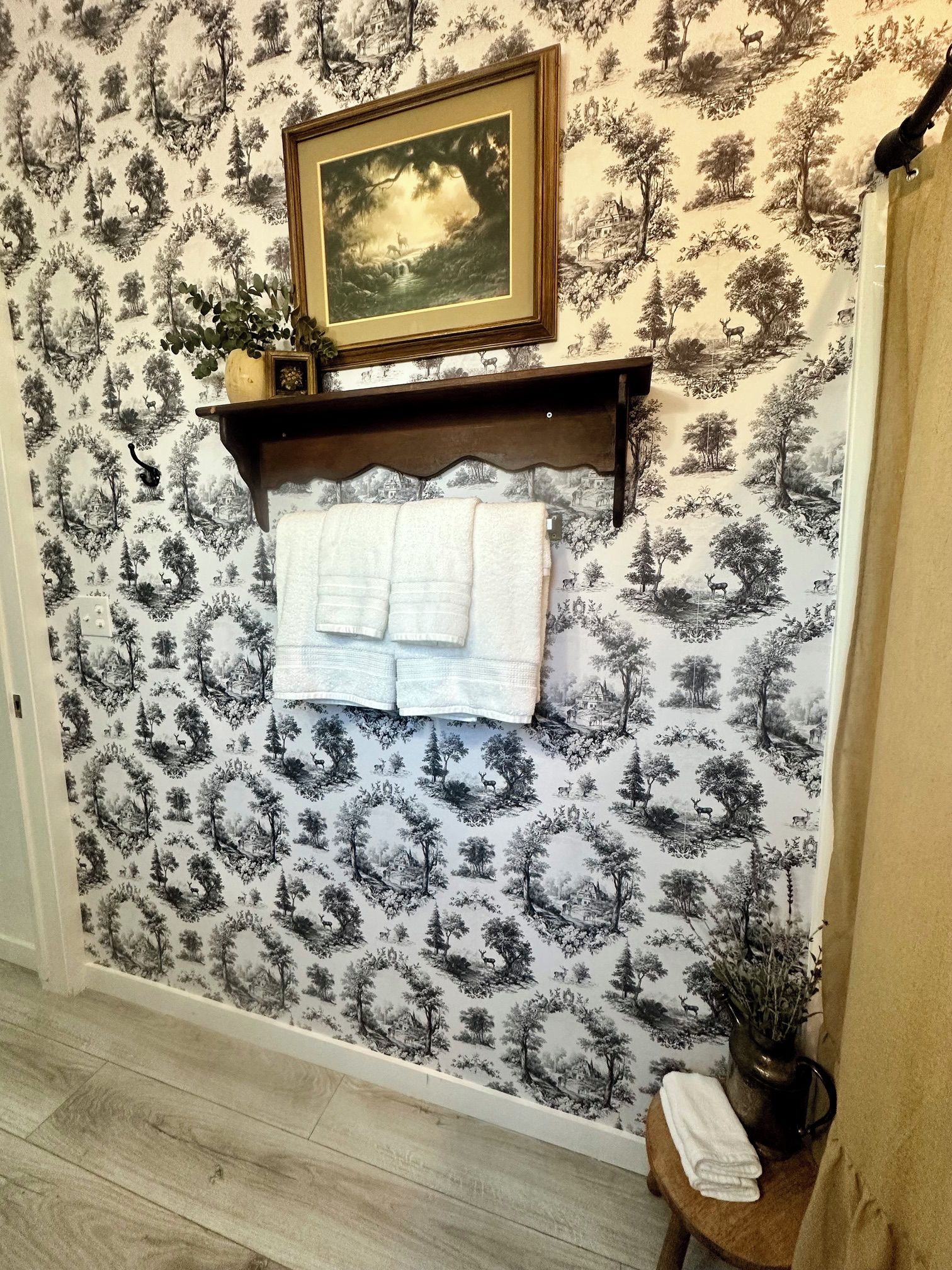 A bathroom with a patterned wallpaper, a framed picture above a towel rack, and a small table with a towel.