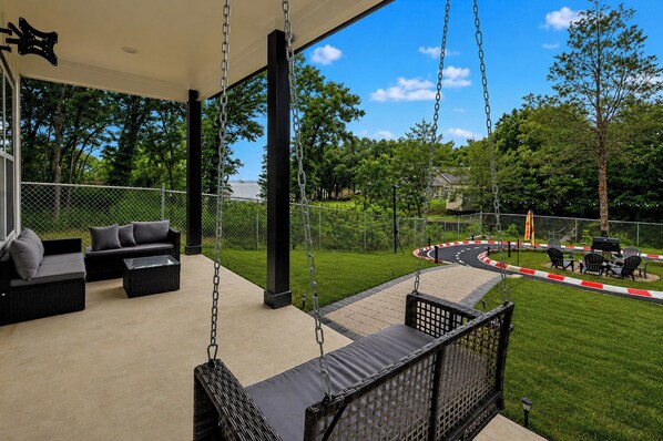 A covered outdoor seating area with swings, a view of a landscaped park, and a fenced pathway.