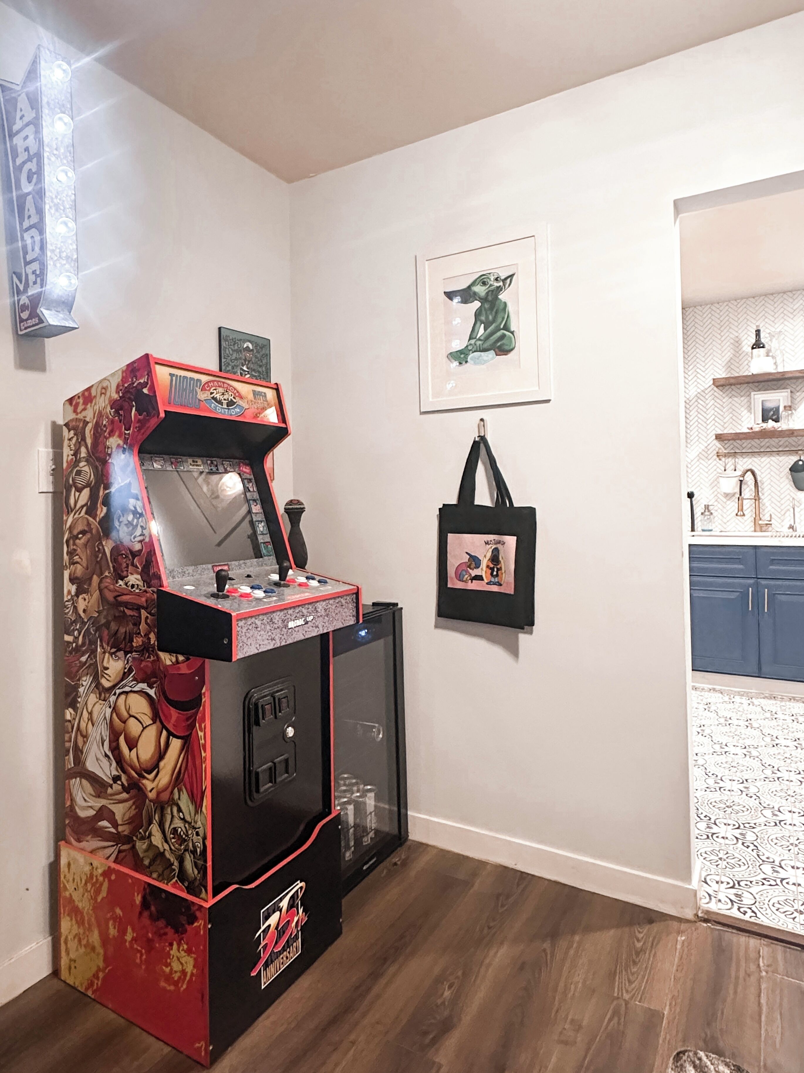 Ein Zimmer mit Spielautomaten, einem gerahmten Charakterbild, einer Wandtasche und einer Küchenzeile mit Regalen.