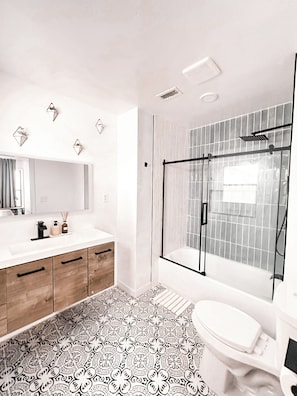 A modern bathroom with a glass shower enclosure, a white sink, and patterned floor tiles. - Quiet Lakewood Gem. A Charming 3-bedroom Home away from Home (Lakewood)