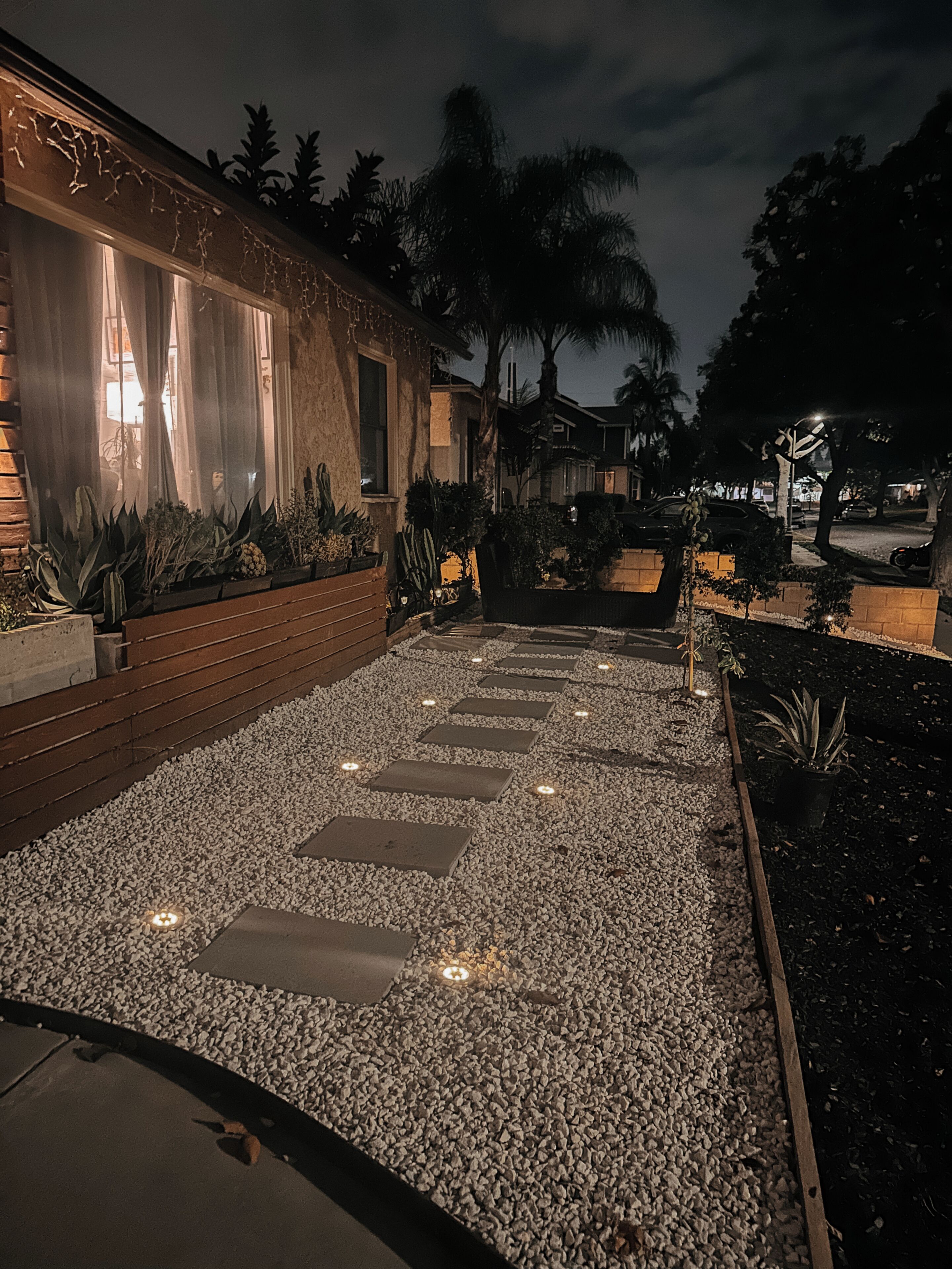 A nighttime outdoor pathway with stepping stones, surrounded by plants and a building with illuminated windows.