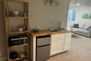 Eine kompakte Küche mit Mikrowelle, Kühlschrank und Spüle. Regale mit Geschirr und ein kleiner Tisch mit Lampe sind vorhanden. Im Hintergrund ist eine Couch zu sehen.