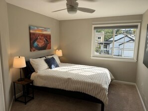 A bedroom with a bed, two bedside lamps, a ceiling fan, a window with a view, and a painting on the wall. - Eco-friendly 4-bedroom Louisville Sanctuary Near Trails (Louisville)
