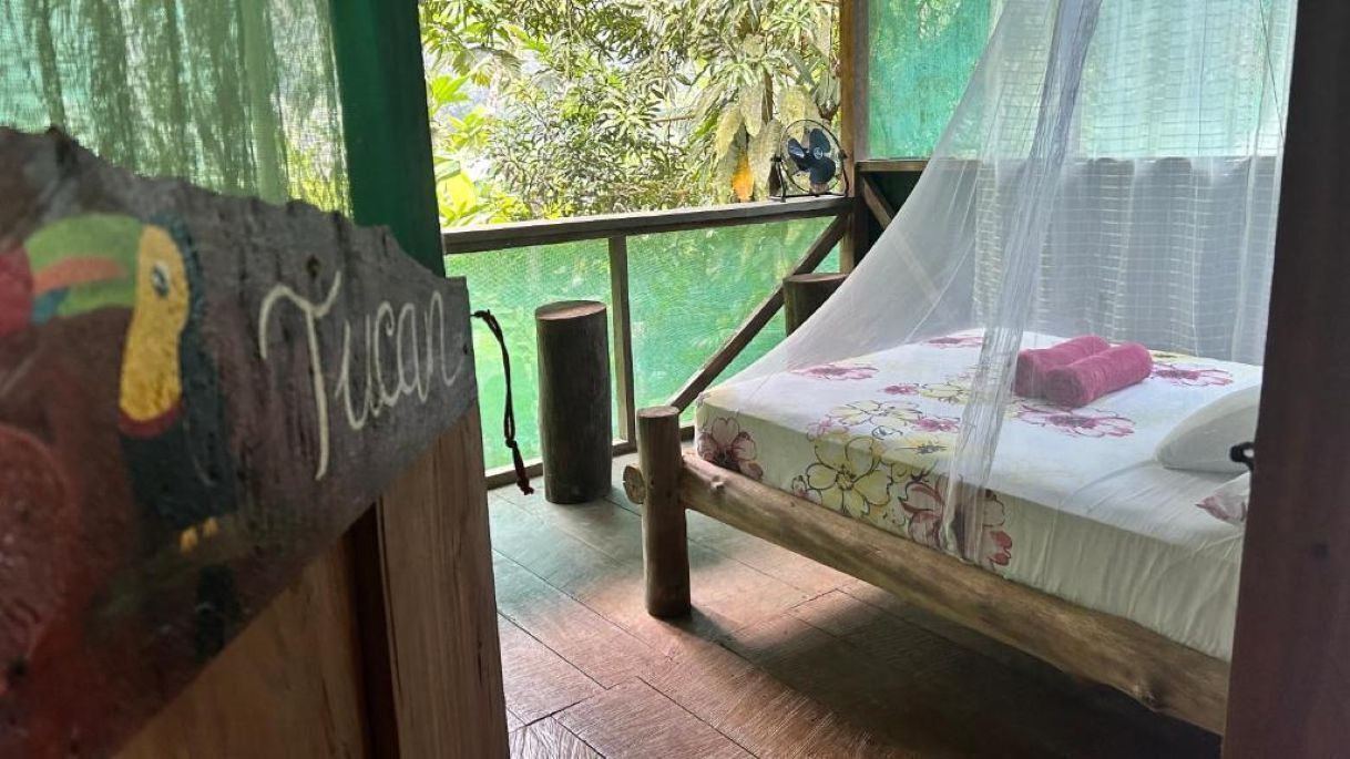 A wooden bed with a floral bedspread and a mosquito net, a wooden bench, and a rustic wall with a painted bird.