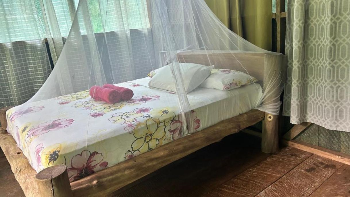 A single bed with a floral bedspread and a red towel, a wooden frame, and a mosquito net.