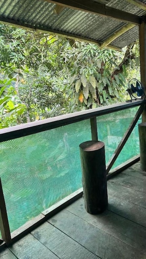 Un espace extérieur abrité doté d’un plancher en bois, d’une rambarde en métal et offrant une vue sur une étendue d’eau à travers un écran en treillis métallique.