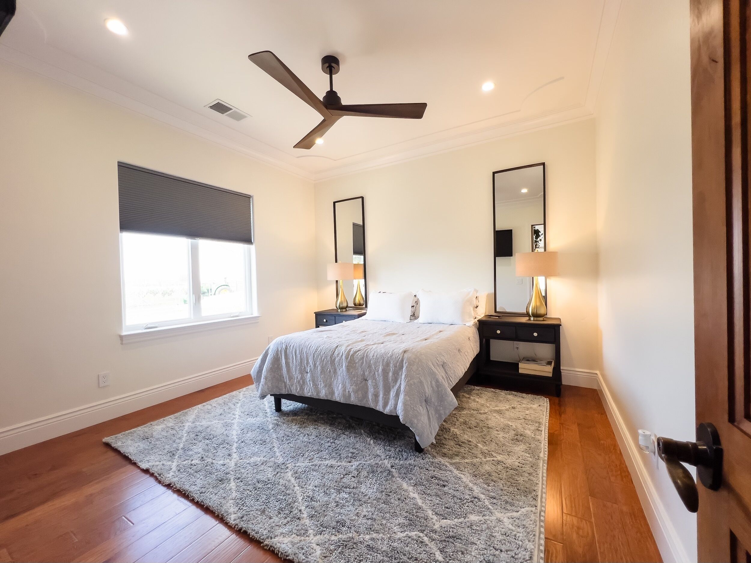 A bedroom with a bed, bedside tables, a ceiling fan, a window with blinds, and a door.