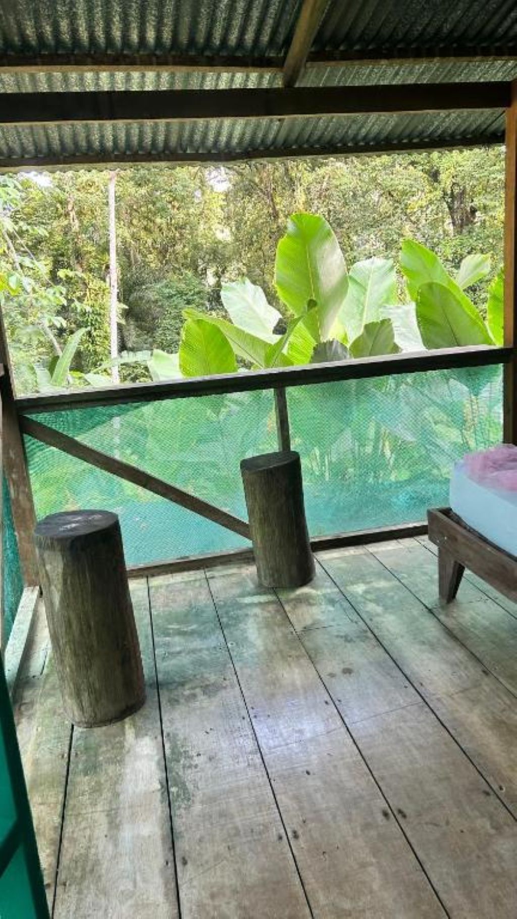 Una terraza de madera con una malla, dos taburetes de madera y una cama con un cojín rosa.