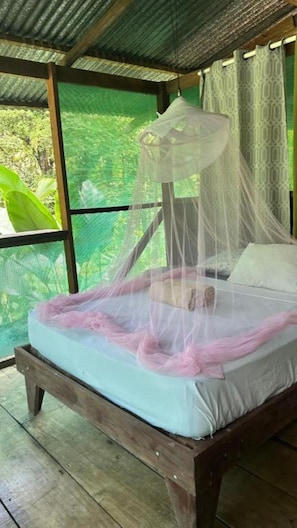 Un letto singolo con zanzariera, struttura in legno e vista sul verde attraverso una finestra a rete.