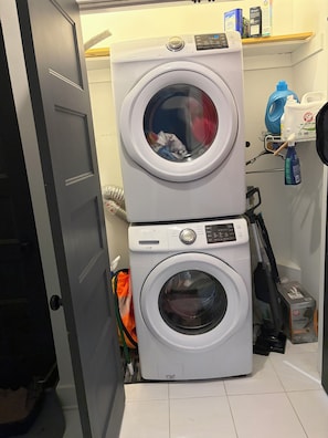 Uma área de lavanderia com duas máquinas de lavar empilhadas, uma prateleira com produtos de limpeza e uma porta que dá para outro cômodo.