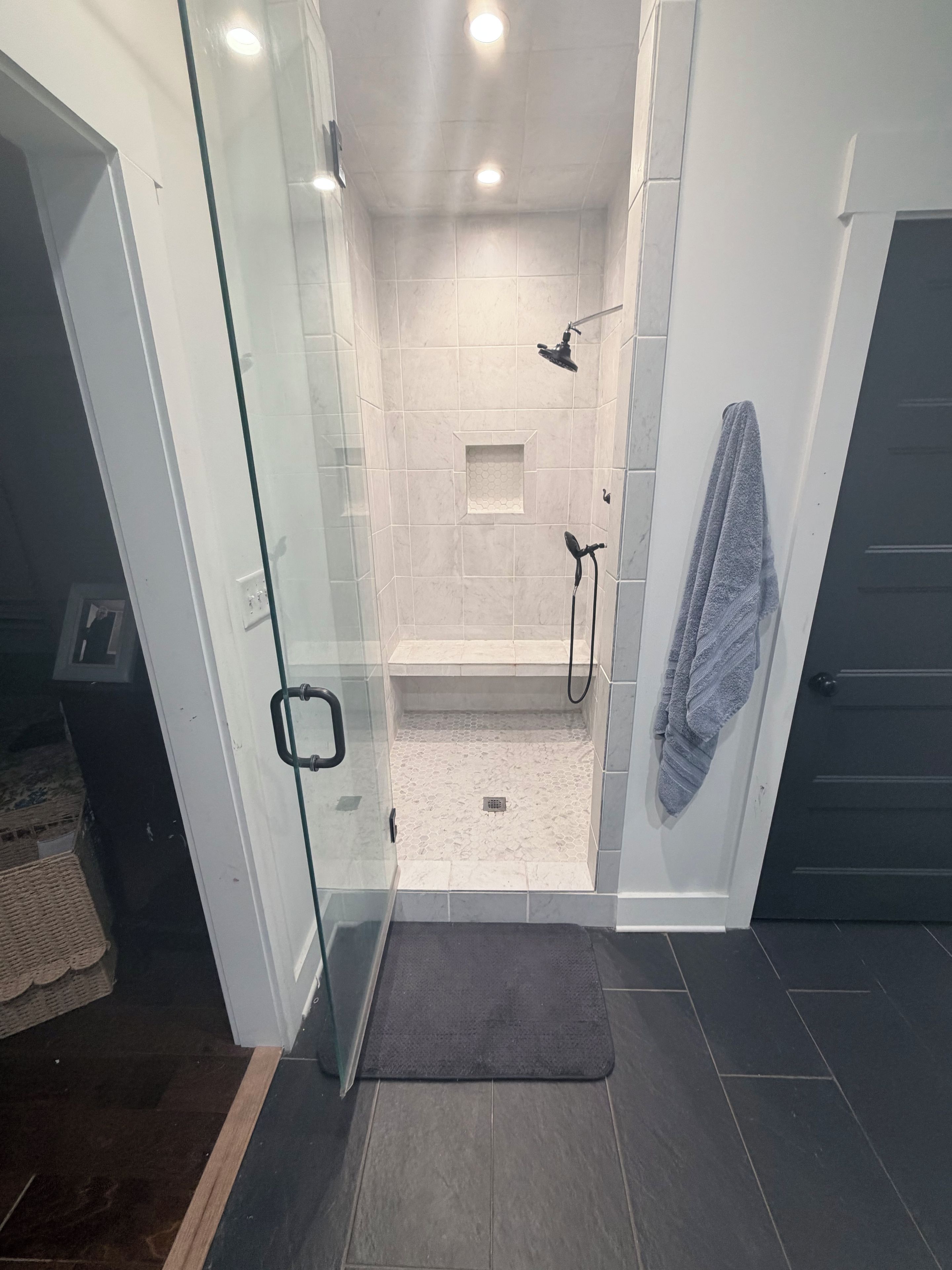 Un espace douche clos par des parois de verre, avec un sol carrelé, une serviette accrochée au mur et un tapis foncé à l’entrée.