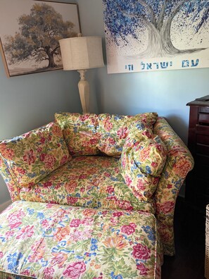 A floral-patterned sofa with a cushion, a lamp, and a framed painting of a tree on the wall.