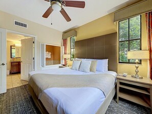 Une chambre d’hôtel avec un grand lit, une table de chevet avec une lampe, une fenêtre avec des rideaux et un ventilateur de plafond.