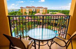 Un balcon doté d’une table et de chaises en verre, offrant une vue sur une piscine et un bâtiment aux toits recouverts de tuiles rouges.