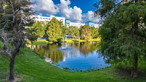 A serene lake with a fountain, surrounded by lush greenery and a building in the background. - 3BR Orlando Resort | Heated Pools | 3 mi Disney (Orlando)