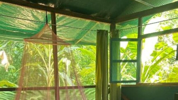 Una cama de madera con mosquitero, un banco y una mesa en una habitación con vistas al verdor.