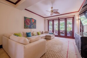 A living room with a sectional sofa, a ceiling fan, a large window with a view of greenery, and a painting on the wall. - Salty Breeze Suite|Seaview Family Residence w/Pool (San Pedro)