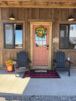 A wooden building with a red door, two Adirondack chairs, a welcome sign, and a wreath.