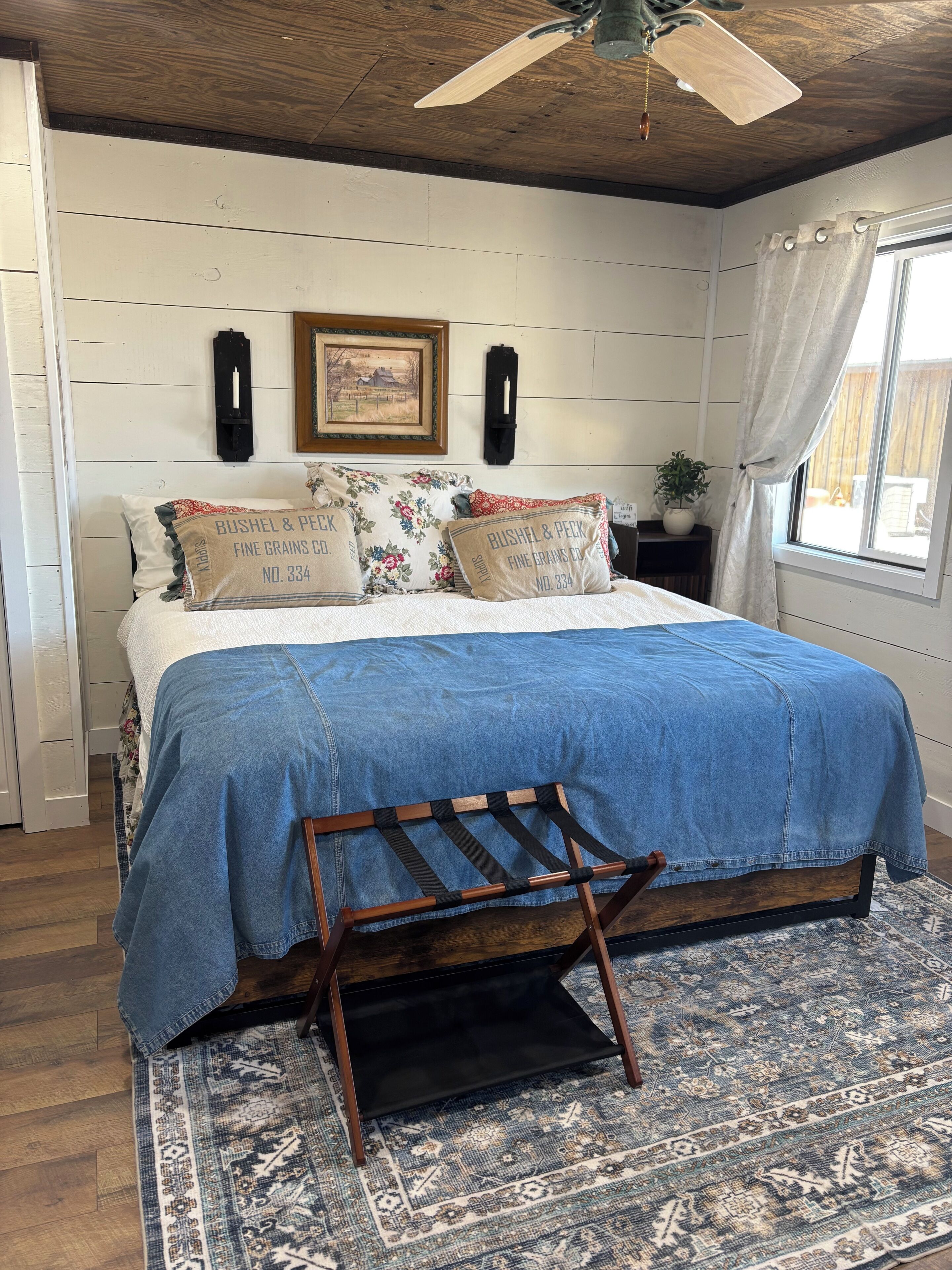 A bedroom with a bed, a wooden bench, a patterned rug, a ceiling fan, and a window with curtains.