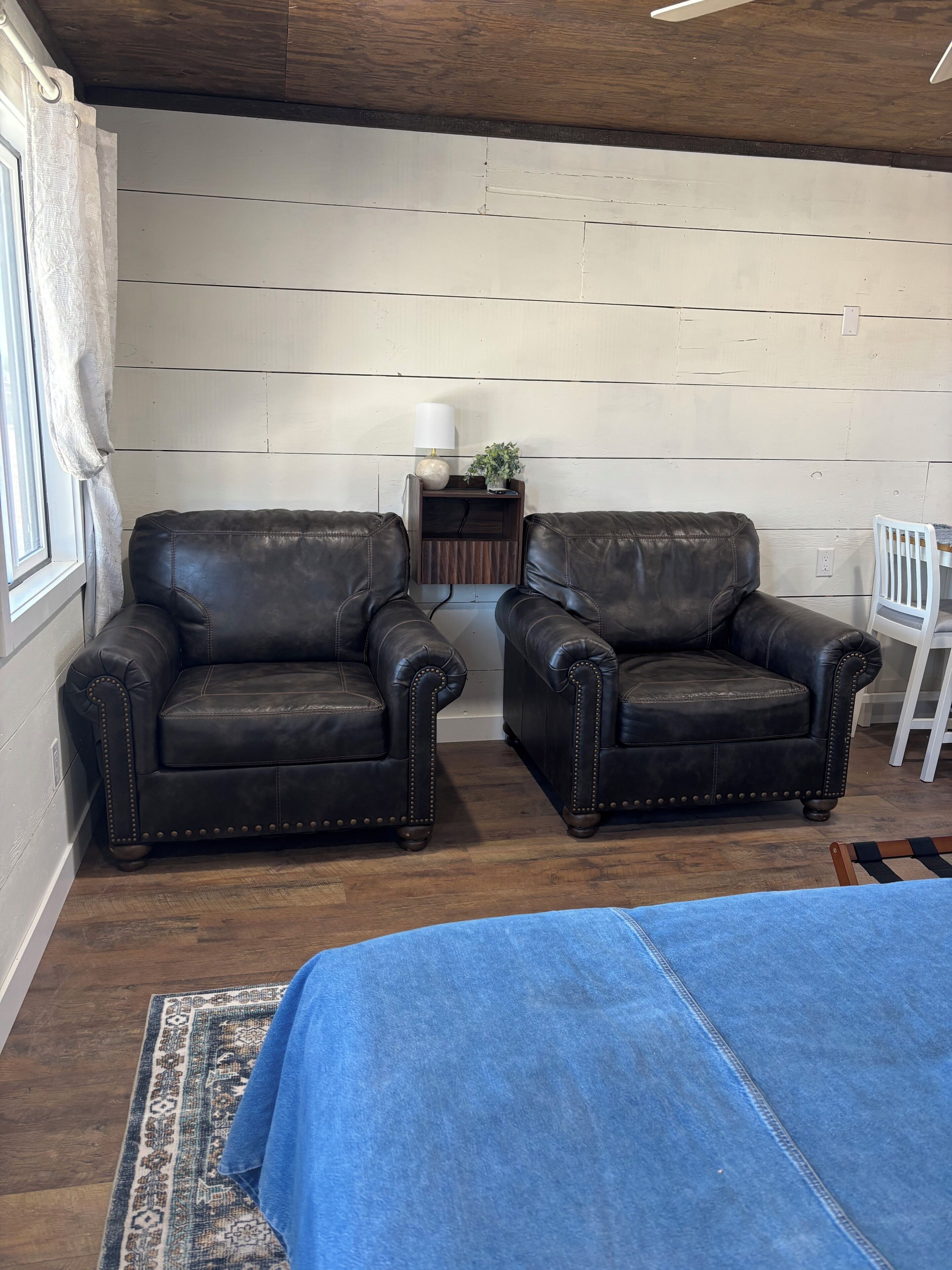A room with two leather armchairs, a small wooden side table with a lamp and plant, a white chair, and a blue bedspread.