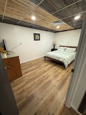 A bedroom with a wooden floor, a bed with a striped bedspread, a nightstand with a lamp, a wall-mounted TV, and a framed picture of a sunflower.