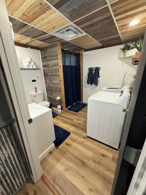 A compact room with a wooden ceiling, a white radiator, a small sink, a toilet, and a washing machine.