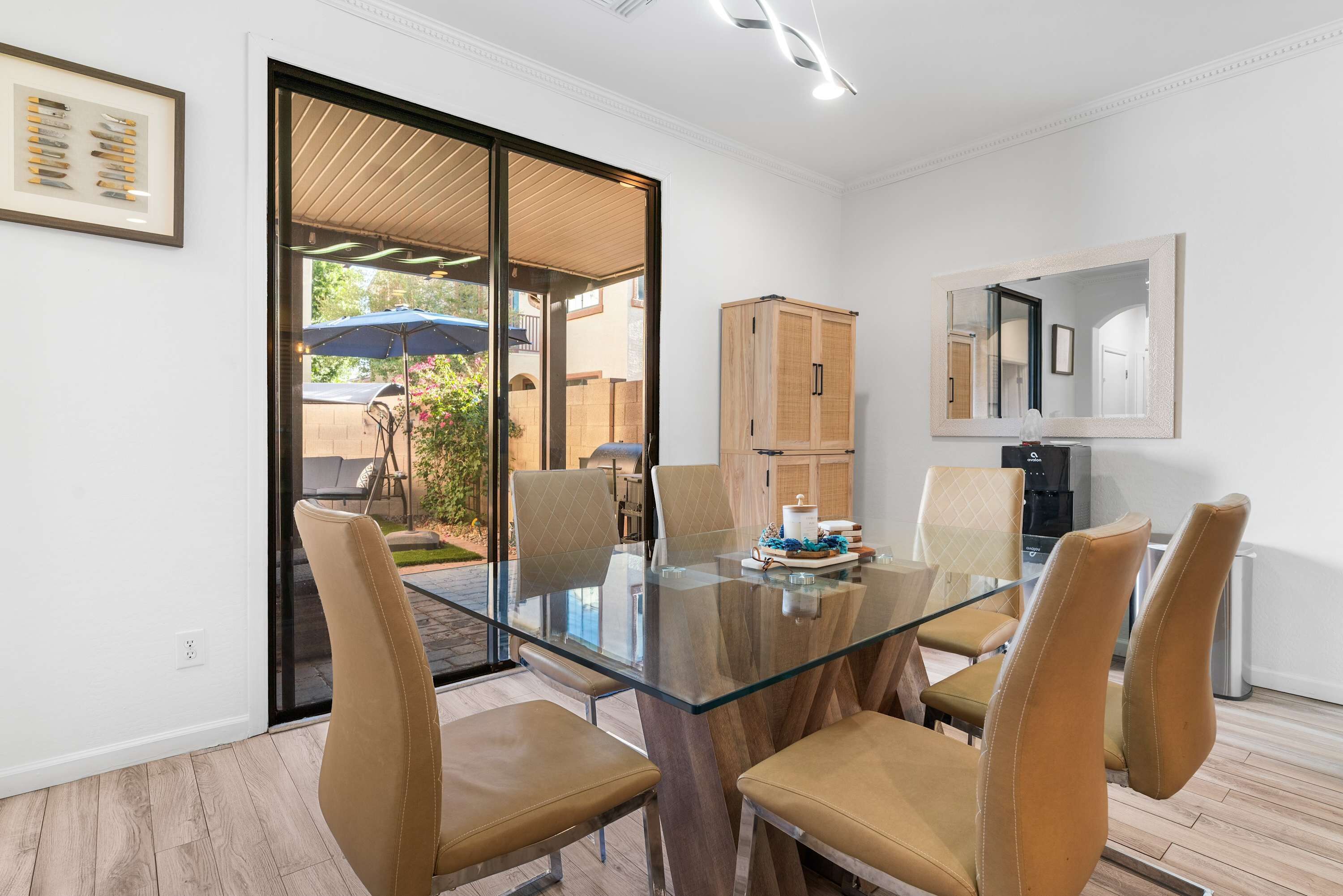 Un comedor moderno con una mesa de vidrio rodeada de sillas de cuero, un armario de madera y una zona de cocina con dispensador de agua.