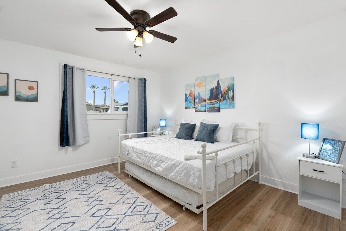 A bedroom with a bed, a ceiling fan, a window with curtains, a nightstand, and wall art.