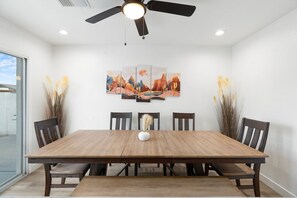 A dining area with a wooden table, six chairs, a ceiling fan, and a wall-mounted painting. - The Desert Palm! Private Oasis! Heated Pool (Glendale)