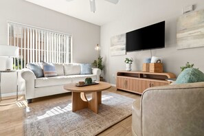 A modern living room with a white sofa, a round coffee table, a wooden cabinet, and a flat-screen TV mounted on the wall. - Fountain Hills Retreat!Pool! Hot Tub! Mountain Views! (Fountain Hills)