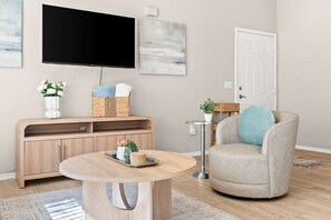 Een woonkamer met een ronde houten salontafel, een beige fauteuil, een flatscreen televisie aan de muur, een geweven mand op een houten dressoir en een deur met een witte omlijsting.