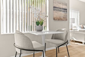 A modern dining area with a white table and two grey upholstered chairs, a potted plant, and a view through large windows with vertical blinds. - Fountain Hills Retreat!Pool! Hot Tub! Mountain Views! (Fountain Hills)