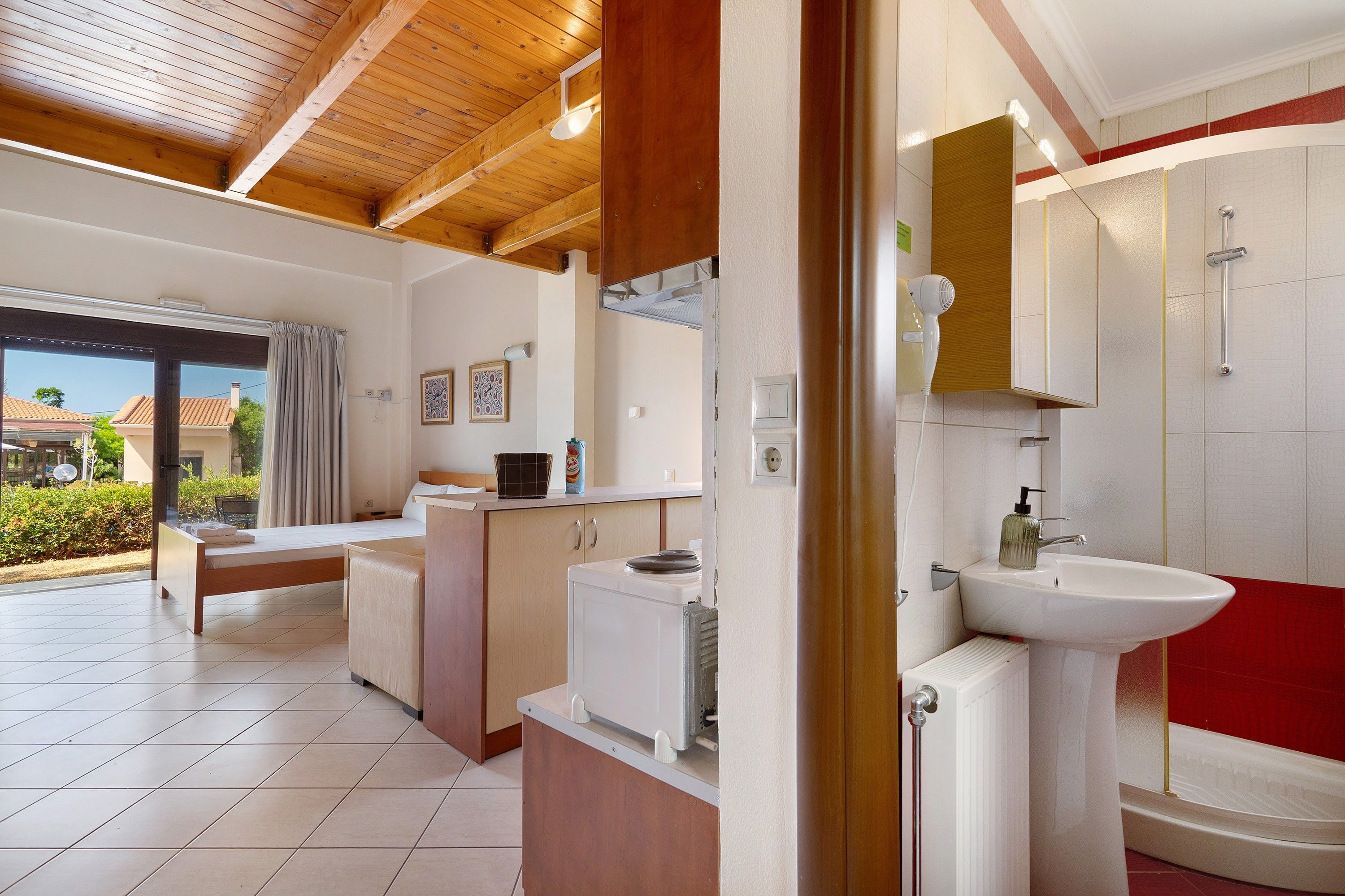 A hotel room with a kitchenette, a large window with curtains, a bathroom with a sink and mirror, and a red accent wall.