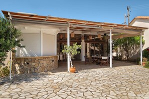 A covered outdoor patio with stone flooring, a potted tree, and a table with chairs. - Villa 'Kalirroe' in Marathiás (Marathiás)