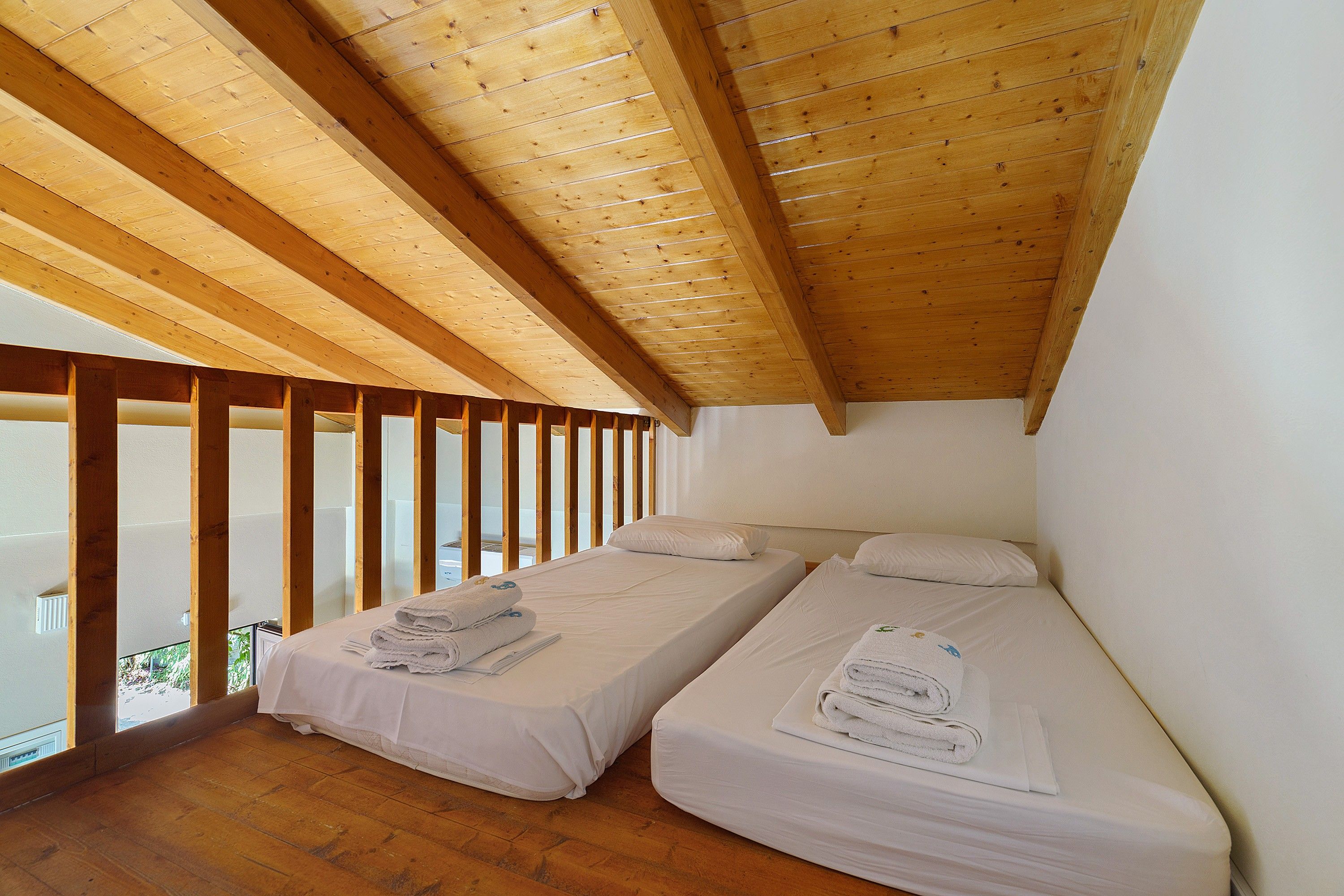 A small, single-room accommodation with two beds, wooden beams on the ceiling, and a balcony with a view.