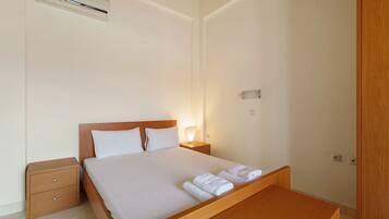 Une chambre d’hôtel avec un lit simple, une table de chevet, une commode, un banc en bois, un appareil de climatisation et une prise électrique murale.