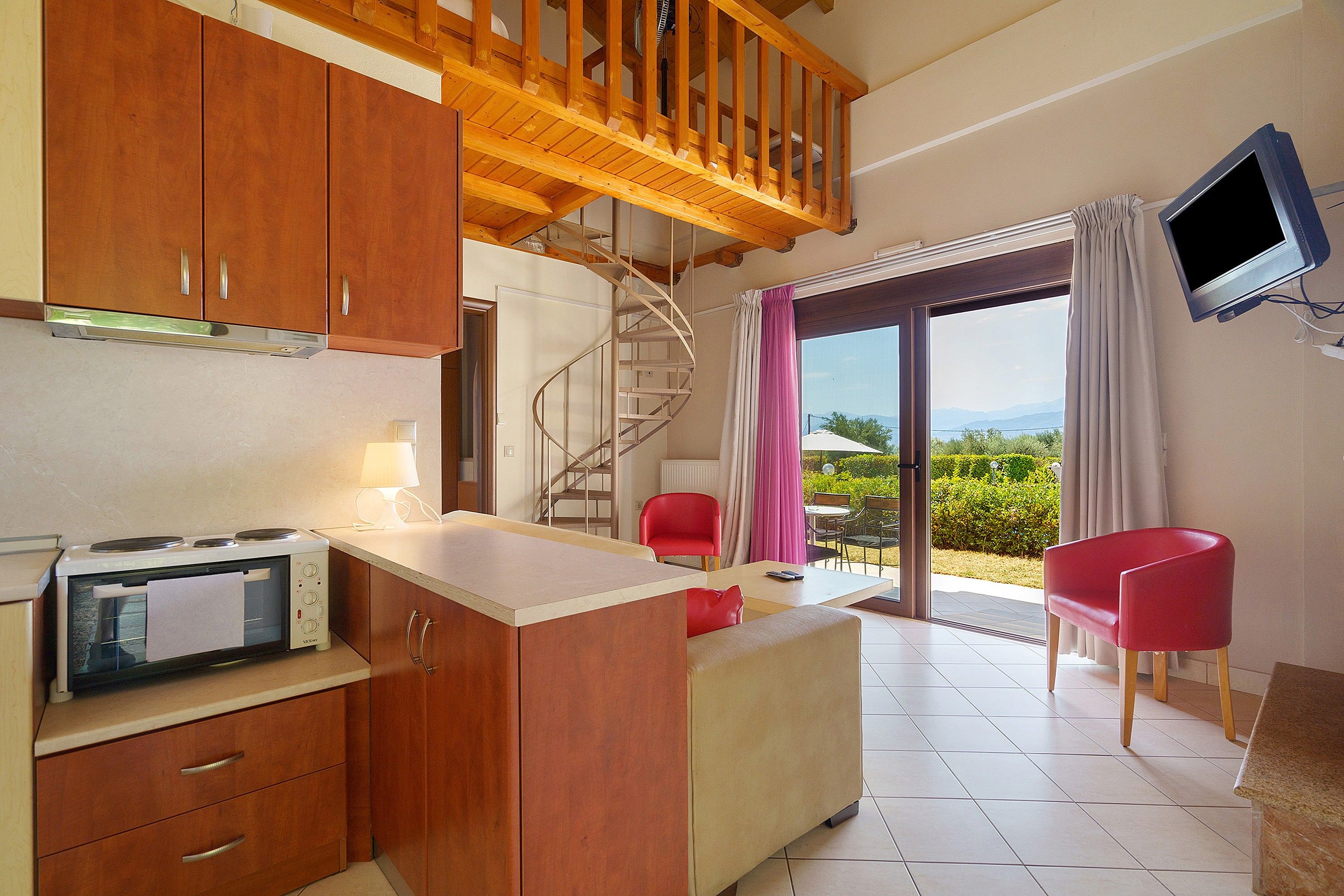 A compact kitchen with wooden cabinets and a built-in stove, a dining area with a table and chairs, and a balcony with a TV and a view of the landscape.