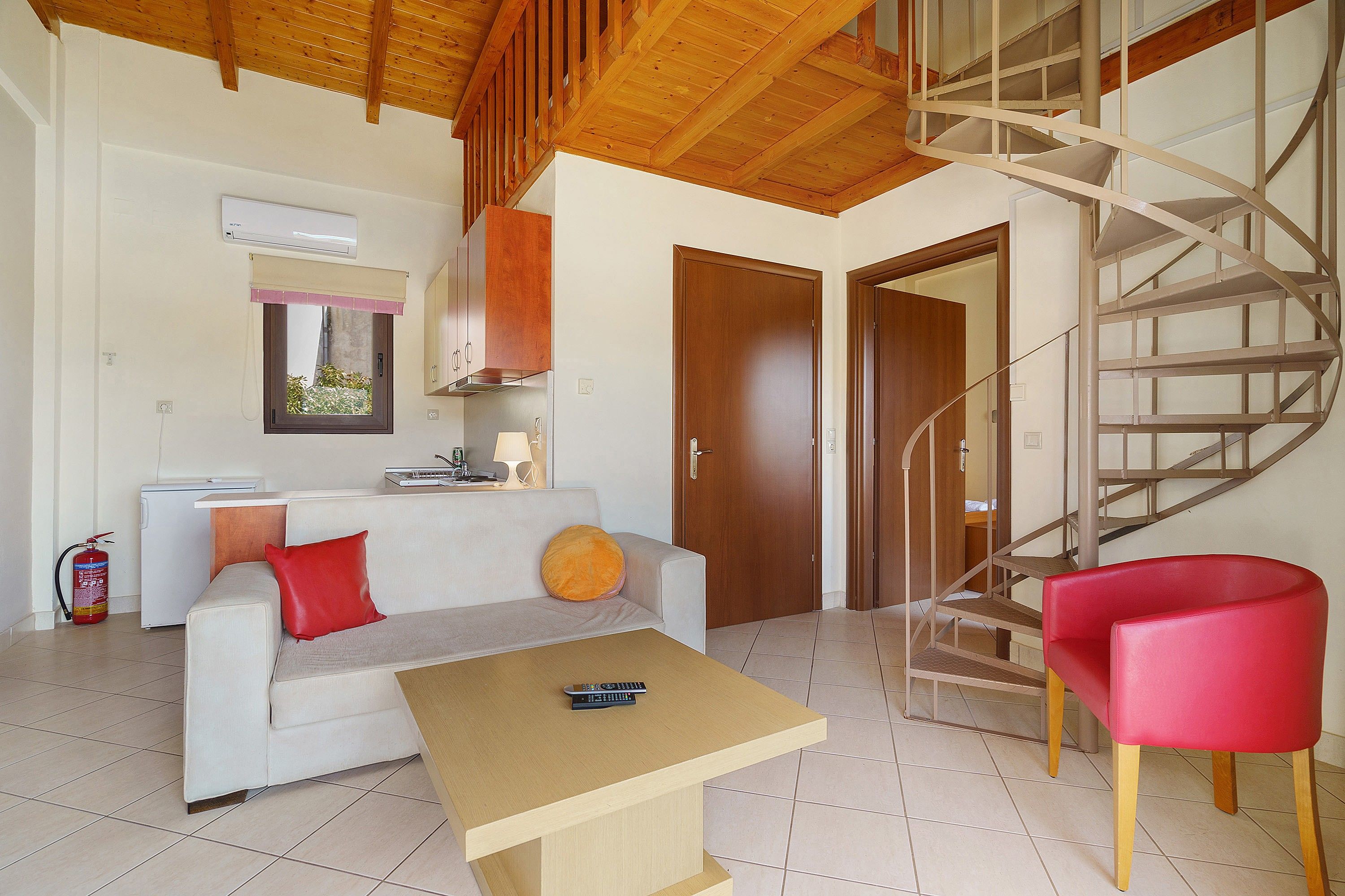 Un espacio habitable compacto con sofá, una mesita, una silla roja, una escalera de madera y una puerta que da a otra habitación.